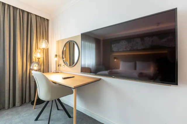 A wall-mounted television above a wooden desk with a grey chair, beside a round mirror and table lamps, with a bed reflected in the dark screen.