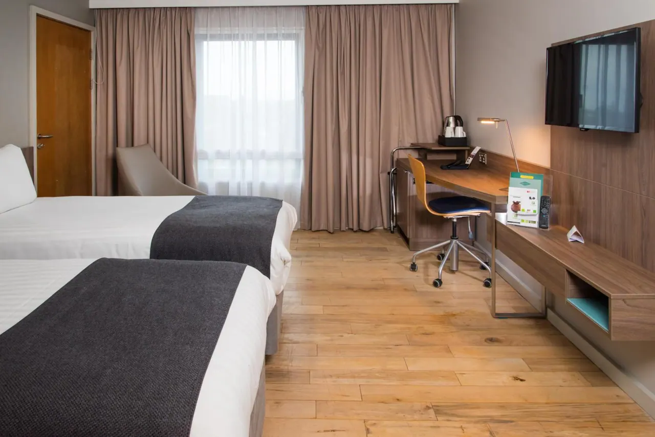A hotel bedroom with two single beds, a desk with a chair, a wall-mounted television and an armchair by the window.
