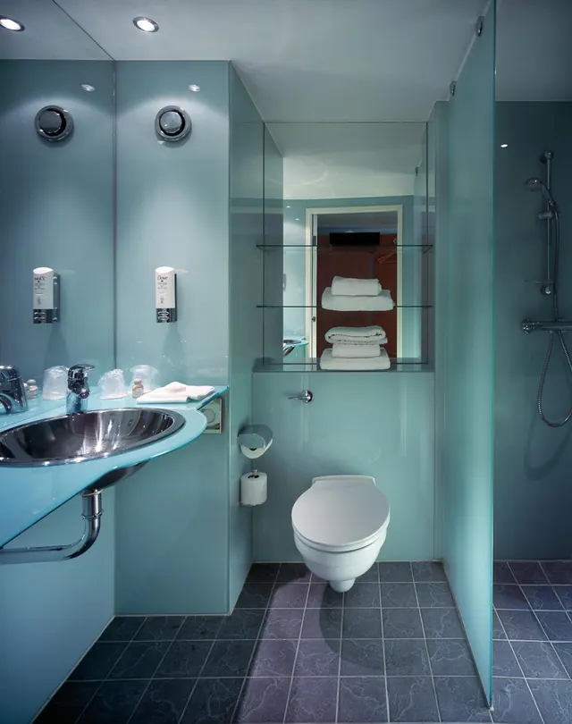 A bathroom with teal glass walls, a wall-mounted toilet, an inset sink, a walk-in shower and folded white towels on glass shelves.