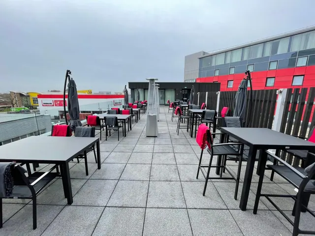 A rooftop terrace with black tables, chairs with red and grey cushions, folded umbrellas and a patio heater, beside a red and grey hotel building.