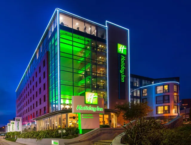 Exterior of a Holiday Inn London West hotel building lit up at dusk, with a green-illuminated glass facade and Holiday Inn signs at street level.