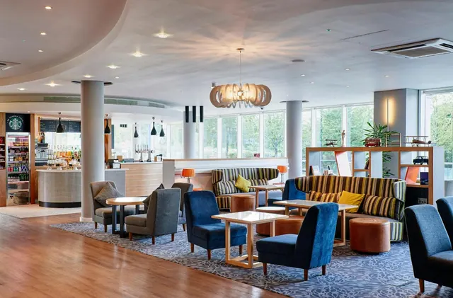 A lounge area with blue armchairs, striped sofas and low wooden tables, with a bar counter on the left and large windows showing trees outside.