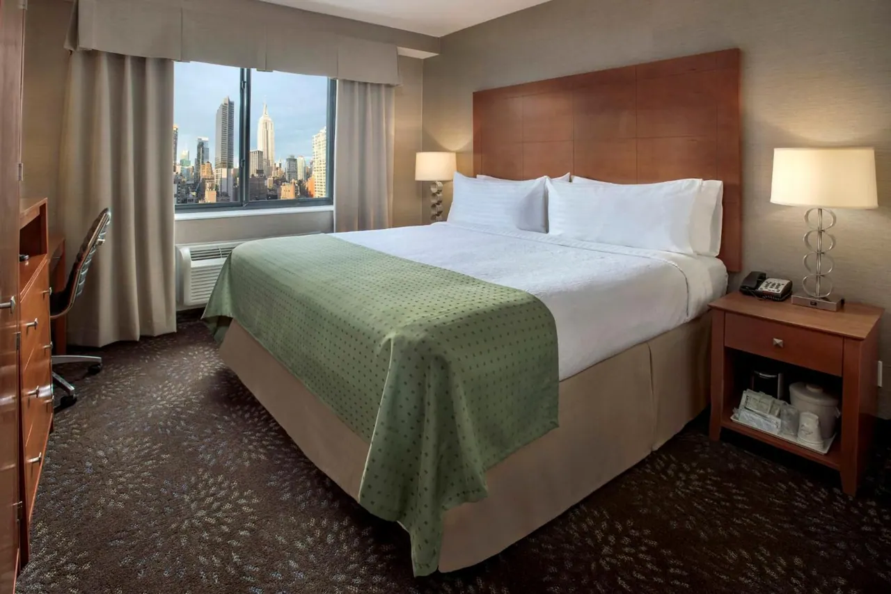 A bedroom with a king bed, a desk and chair by the window, and a view over city buildings including the Empire State Building.