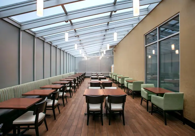 A glass-roofed dining area with wooden tables, upholstered chairs, green armchairs and a banquette, pendant lights, and large windows along the right wall.