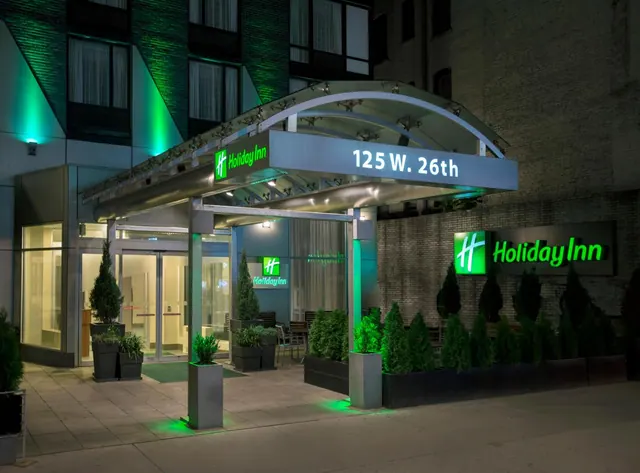 Night view of a hotel entrance under a curved metal canopy, with illuminated green signage and potted evergreens along the facade.