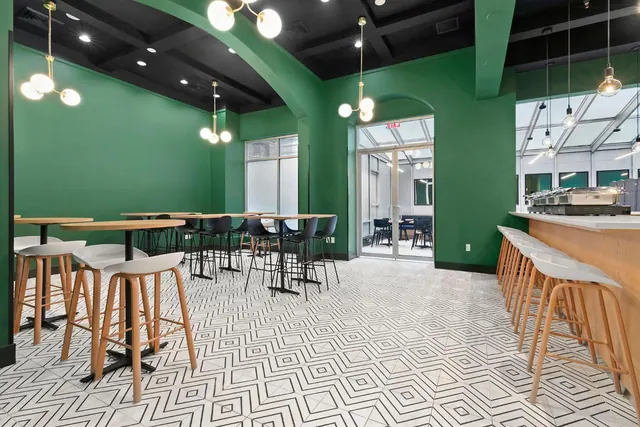 A dining room with green walls, high bar tables and stools, a geometric patterned tile floor, and a counter with chafing dishes on the right.