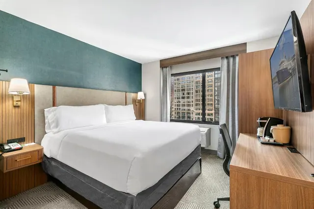 A made king bed with white linens sits against a teal accent wall, beside a wooden desk, a wall-mounted television, and a window looking out onto tall city buildings.