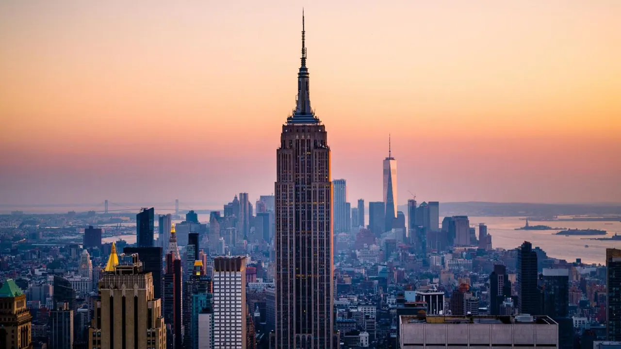 Elevated sunset view over the Manhattan skyline with the Empire State Building at centre and One World Trade Center to the right, with water visible on the right.