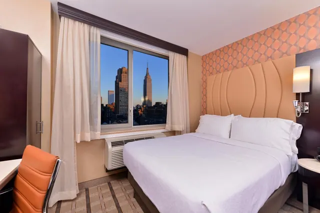 A bedroom with a made king bed, an orange desk chair and a wardrobe, with a window view of the Empire State Building.
