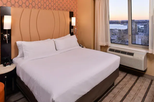 A king bed with white linens and a padded headboard sits beside a window overlooking city buildings and a body of water at dusk.