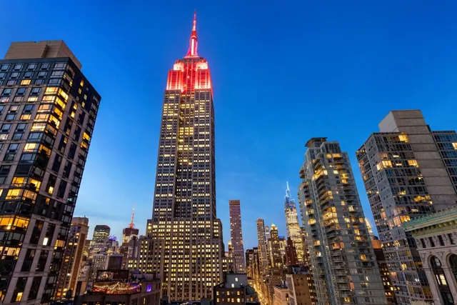 Dusk view of the Empire State Building, lit red at the top, rising above surrounding midtown towers with lit windows.