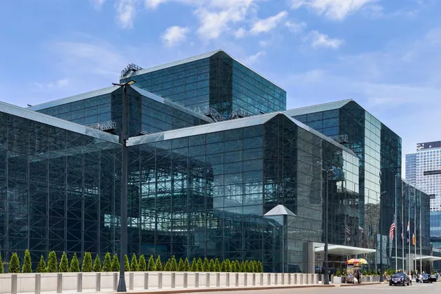Exterior of a large glass-faceted convention center with a street, parked cars and a food cart in front under a partly cloudy blue sky.