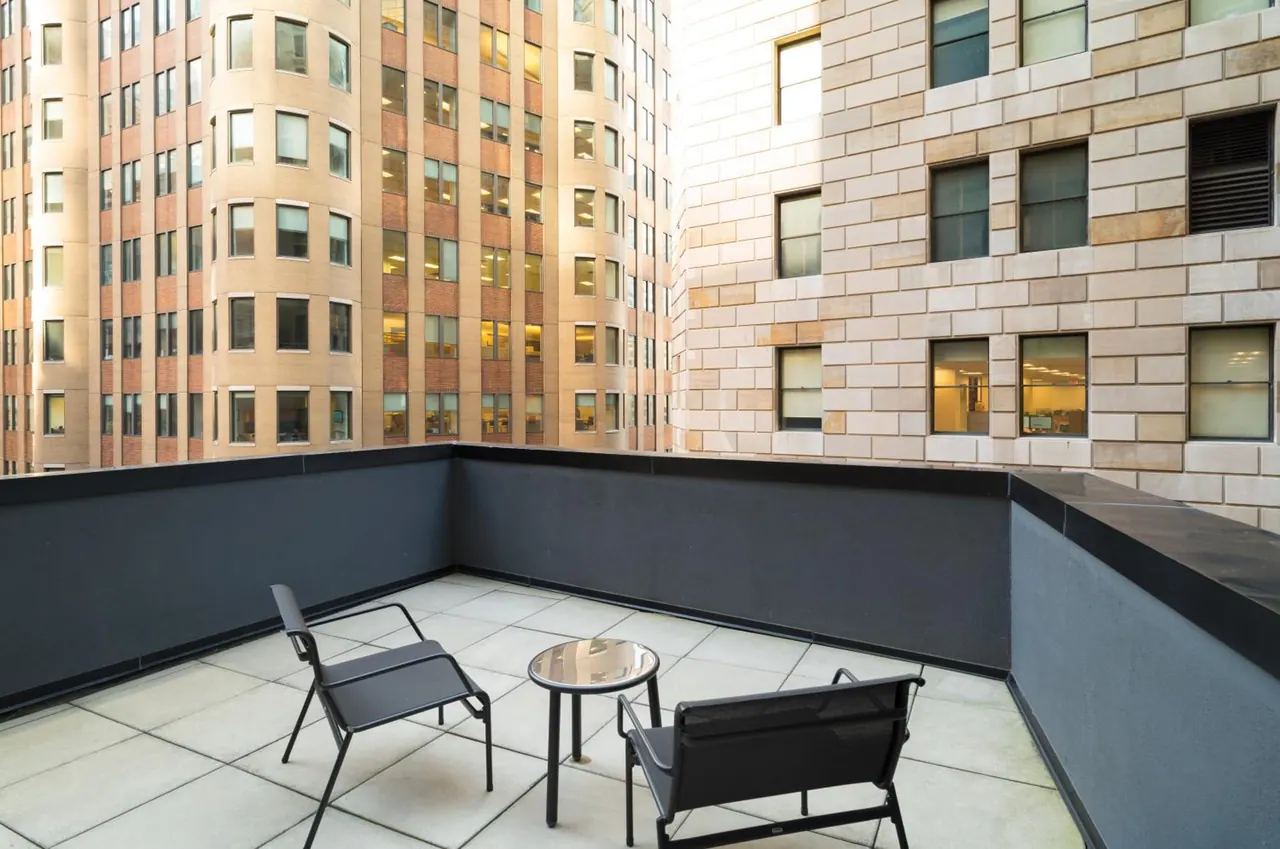 A furnished rooftop terrace with two chairs and a small round table, with office buildings visible beyond the dark parapet walls.