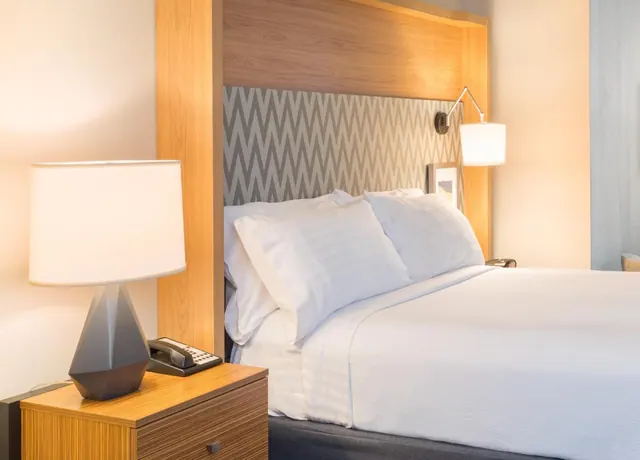 A bedroom with a made bed with white pillows, a wall-mounted reading lamp, and a bedside table holding a lamp and a telephone.