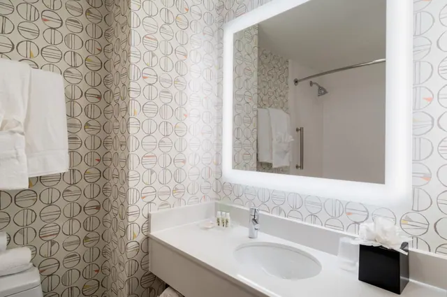 A bathroom with patterned wallpaper, a backlit mirror above a single sink vanity, and a shower stall visible in the reflection.