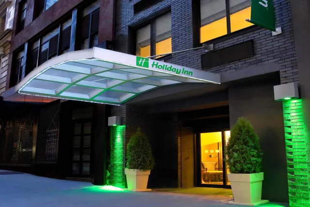 The entrance of a Holiday Inn at dusk, with a glass canopy, green-lit brick pillars and two potted topiary trees beside the open door.