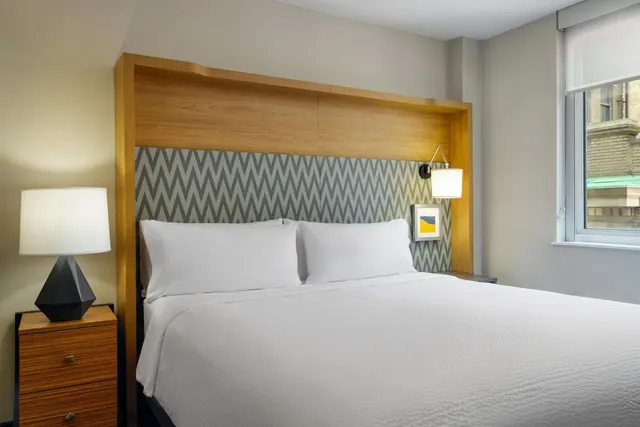 A bedroom with a made king bed, a wooden headboard set against a chevron-patterned wall, bedside lamps, and a window looking onto a brick building.