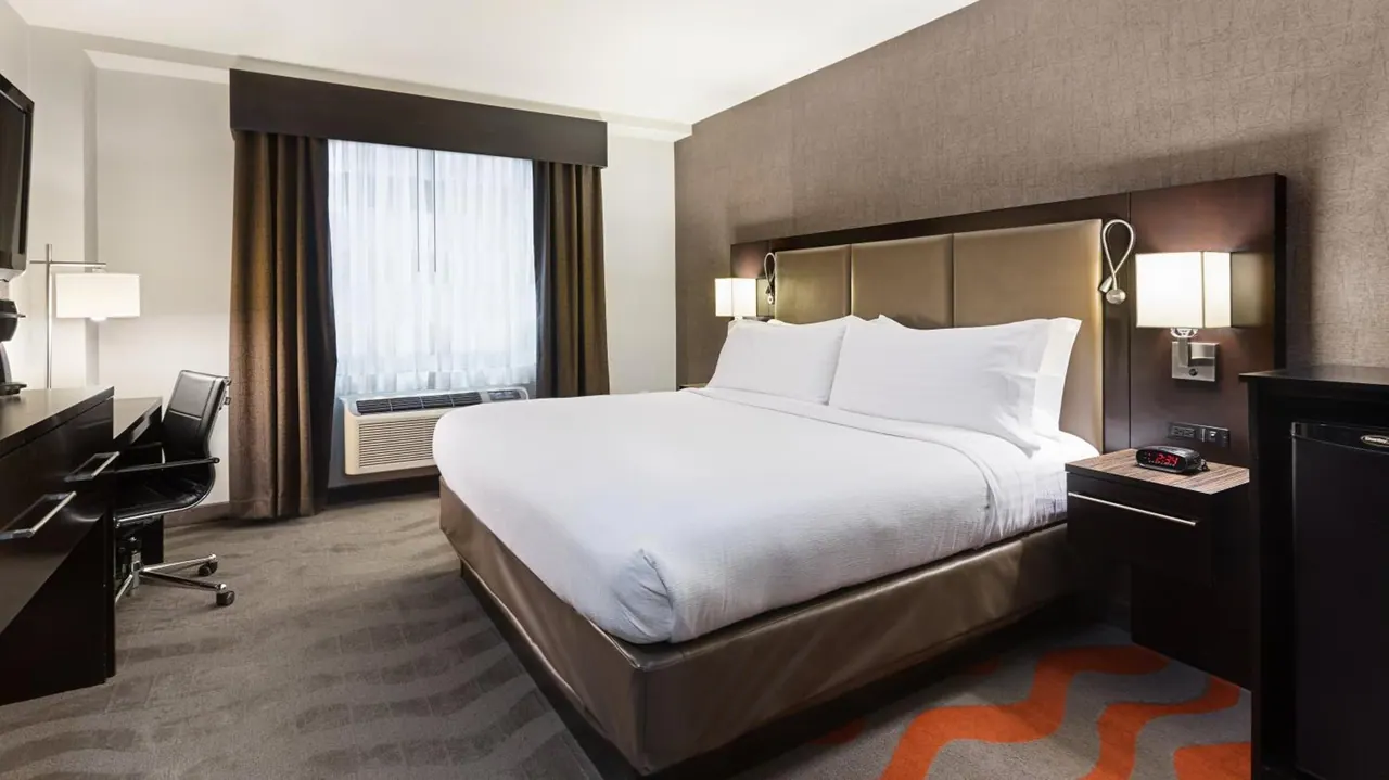 A hotel bedroom with a king bed, a work desk and chair, a wall-mounted television, and a window with an air conditioning unit beneath it.