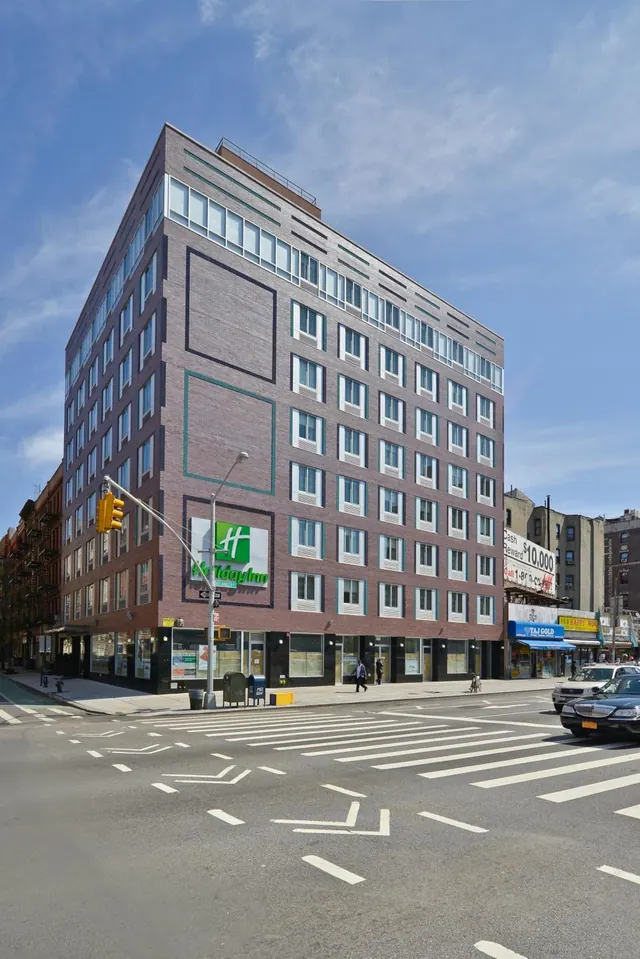 Exterior of a brick Holiday Inn Express building on a city corner, with a crosswalk, parked cars and a few pedestrians on the street.