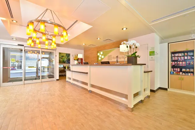 A hotel lobby with a wooden reception counter, an orange ring chandelier, glass entrance doors and a light wood-effect floor.