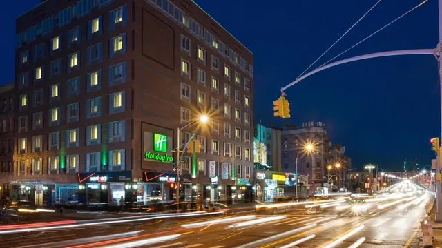 Night exterior of a brick Holiday Inn building beside a busy street, with traffic light trails and a traffic signal in view.