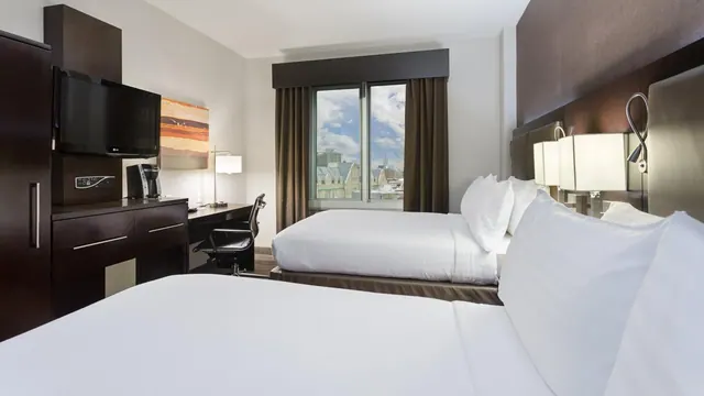 A hotel room with two made beds, a wall-mounted television, a desk with a chair and coffee machine, and a window showing city buildings and sky.