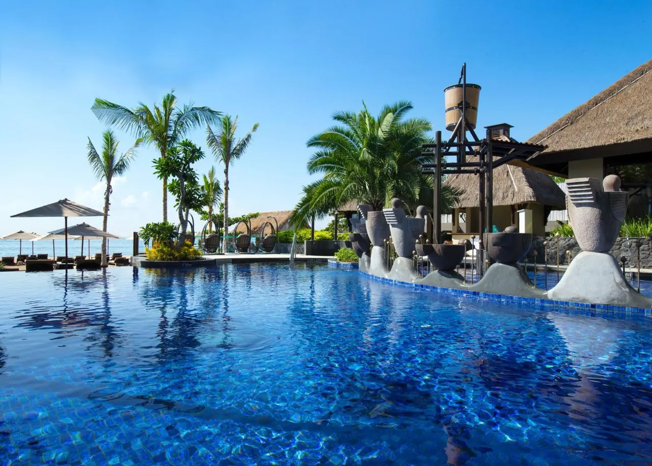 A large blue-tiled swimming pool with sun loungers and umbrellas, palm trees, stone water sculptures, and the sea visible in the background.