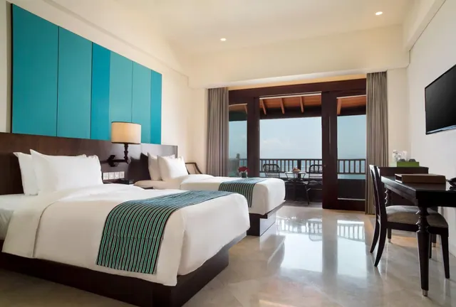 A bedroom with two beds, a work desk, a wall-mounted television, and glass doors opening onto a balcony with a view of the sea.