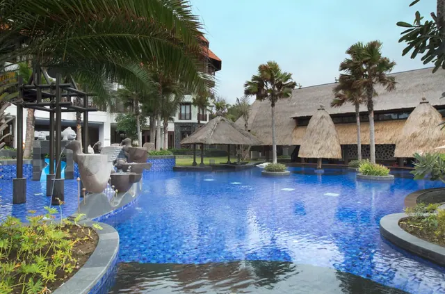 A large blue-tiled swimming pool with a sculptural fountain on the left, with thatched pavilions and palm trees beyond.