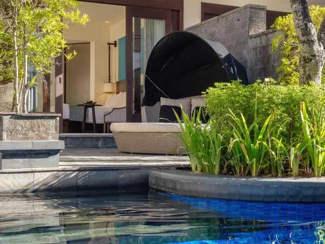 A pool edge beside a terrace with a canopy daybed, with a bedroom visible through open glass doors.