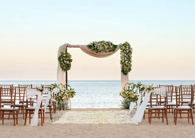 A beach wedding ceremony setup with a draped, flower-covered arch and rows of chairs on the sand, facing the sea.