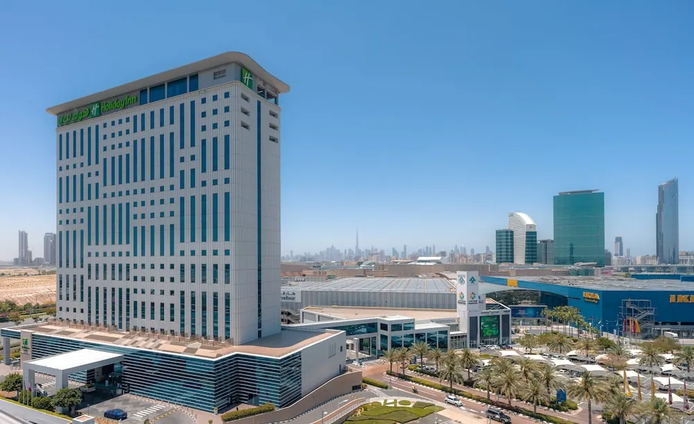 Exterior of a tall white Holiday Inn hotel tower beside a shopping area with palm trees, parked cars and the Dubai skyline in the distance under a clear blue sky.