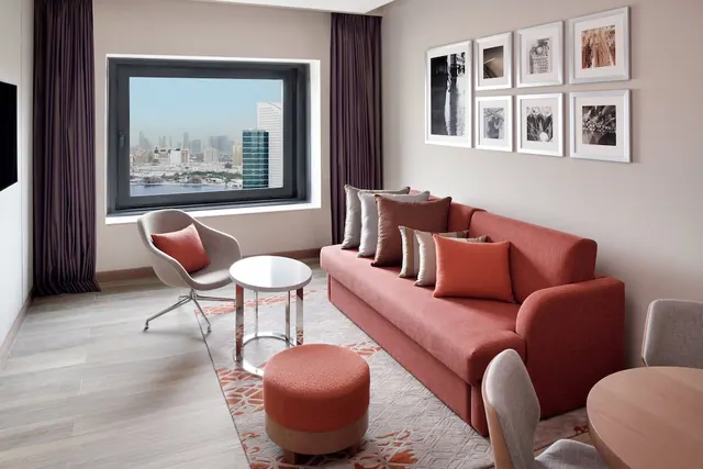 A living area with a red sofa, round side table and ottoman, and framed black and white prints on the wall, with a window looking over a city skyline and water.