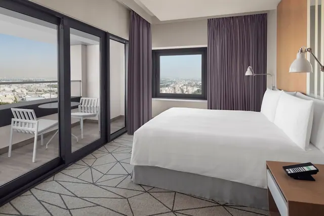A hotel bedroom with a made king bed, purple curtains, and sliding glass doors opening onto a balcony with white chairs and a view over a hazy city.