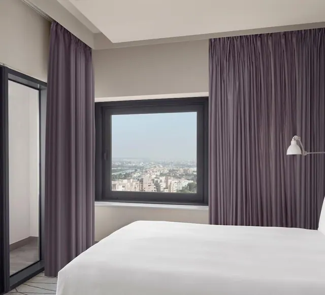 A bedroom with a made white bed, purple curtains, a wall lamp, and a window looking out over a hazy city of low-rise buildings.