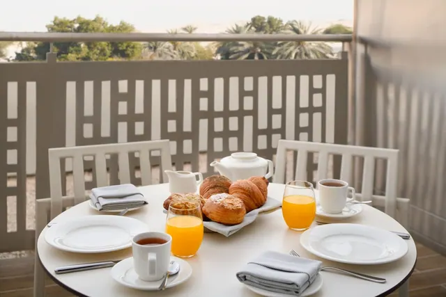 A breakfast table with pastries, orange juice, tea and a teapot sits on a balcony with a view of trees and palm trees beyond the railing.