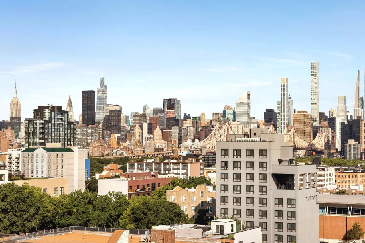 Elevated view over neighbouring rooftops and buildings toward the Manhattan skyline, with the Empire State Building and the Queensboro Bridge visible under a clear sky.