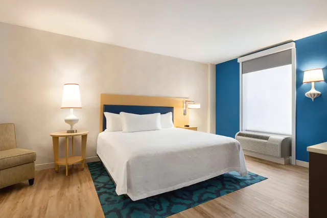 A bedroom with a made king bed, a bedside lamp, an upholstered armchair, a blue accent wall and a wall-mounted air conditioning unit under the window.