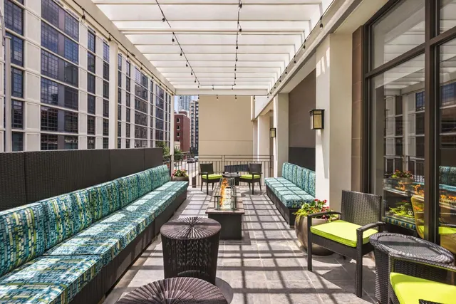 A covered outdoor terrace with long cushioned benches, wicker chairs, a fire pit table and string lights, with city buildings visible beyond the railing.