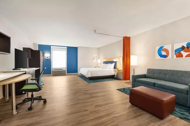 A hotel room with a king bed, a grey sofa, a desk with a chair, and a wall-mounted television, with a bright window at the back.