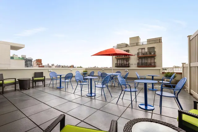 A rooftop terrace with blue metal chairs and round tables, a red umbrella, green-cushioned lounge chairs, and a neighbouring building with distant city buildings beyond.