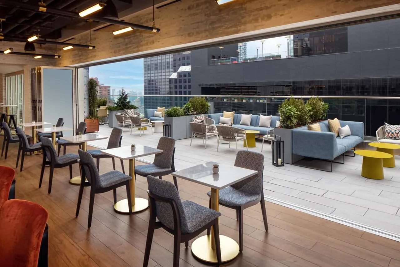 A rooftop terrace with dining tables, blue sofas and yellow side tables, with tall city buildings visible beyond the glass railing.