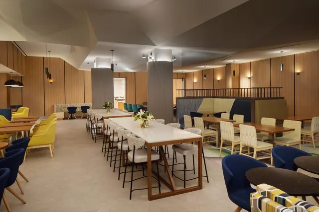 A dining area with long wooden-topped tables and bar stools, wood-panelled walls, upholstered booths, and round dark tables with blue armchairs.