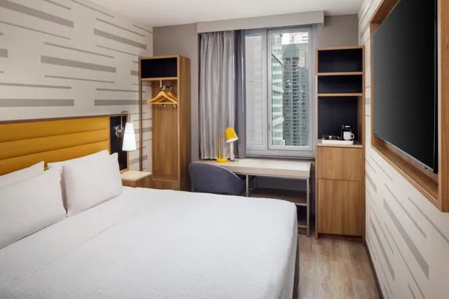 A bedroom with a king bed, a wall-mounted television, a wardrobe, a desk with a chair, and a window showing tall city buildings.