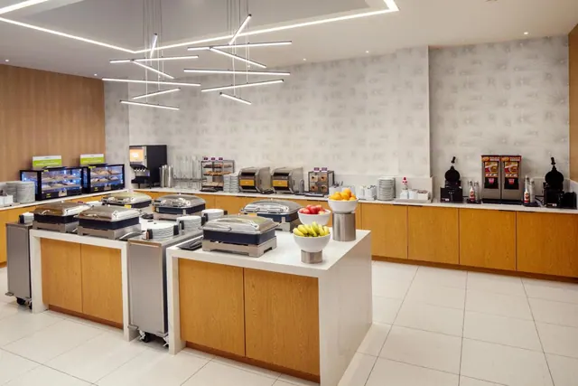 A hotel breakfast buffet room with covered chafing dishes, fruit bowls and a coffee station along a wood-fronted counter under linear ceiling lights.