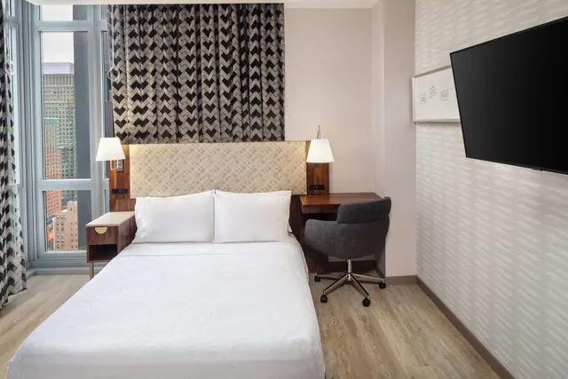 A bedroom with a made double bed, a desk with a chair, a wall-mounted television and a window looking out onto city buildings.