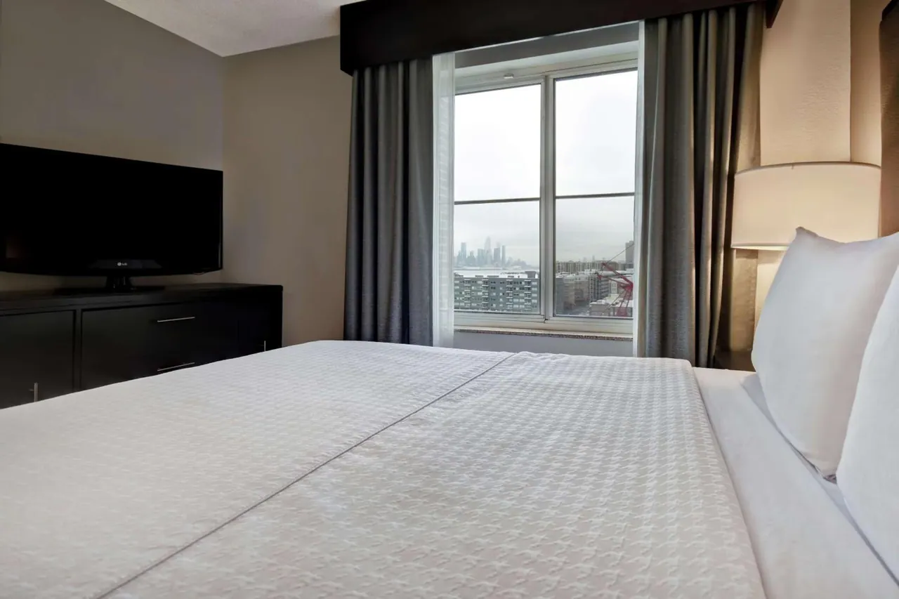 A bedroom with a made king bed, a flat-screen television on a dark dresser, and a window showing a distant city skyline across the water.