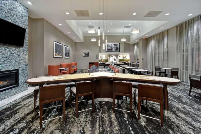 A large curved bar-height table with stools sits in a lounge area with a wall-mounted television, a stone fireplace, orange armchairs, and a breakfast counter in the background.