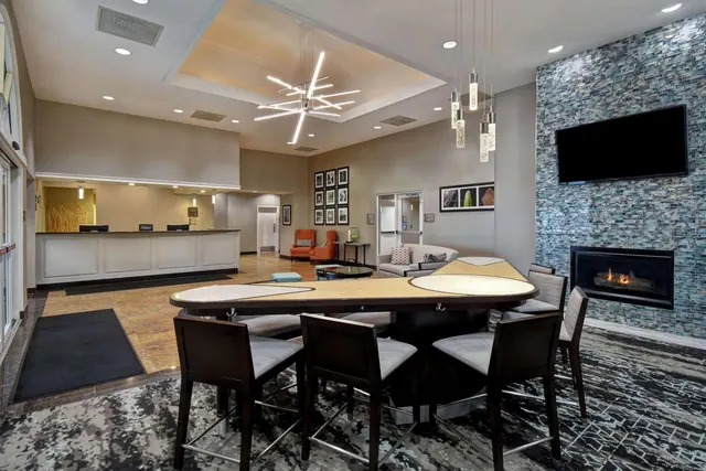 A hotel lobby with a reception desk, a high-top table with bar stools, a seating area, and a wall-mounted television above a mosaic-tiled fireplace.