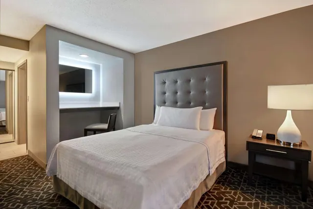 A bedroom with a made double bed, a tufted grey headboard, a bedside lamp and a desk with a lit mirror.